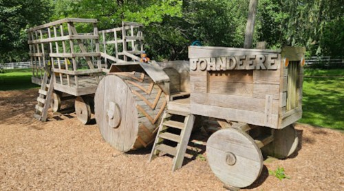 A wooden playground structure designed like a tractor and trailer, set in a grassy area with trees.