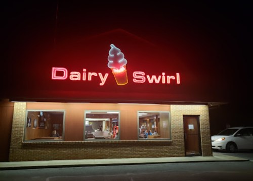 A brightly lit sign reading "Dairy Swirl" with an ice cream cone, outside a cozy ice cream shop at night.