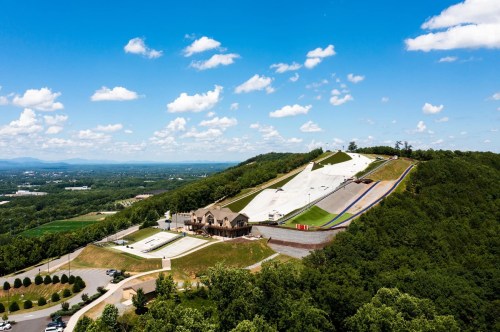 Aerial view of a ski slope with a lodge, surrounded by green hills and a blue sky with scattered clouds.