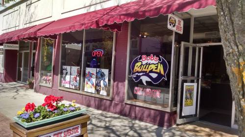 Exterior of Bob's Bullpen comic store, featuring large windows with colorful displays and a flower planter in front.