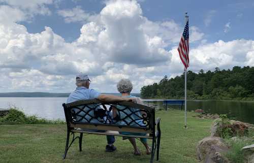 An elderly couple sits on a bench by a lake, enjoying the view with an American flag in the background.