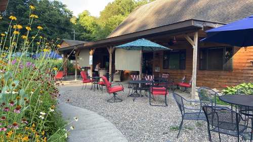 A cozy outdoor seating area with red chairs, tables, and colorful flowers, set against a rustic wooden building.