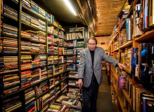 A man in a gray blazer stands in a narrow aisle surrounded by tall bookshelves filled with books.
