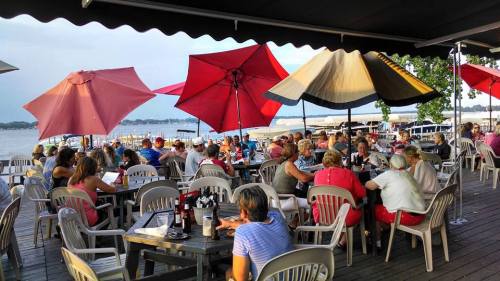 A lively outdoor dining area by the water, with patrons under large umbrellas enjoying food and drinks.