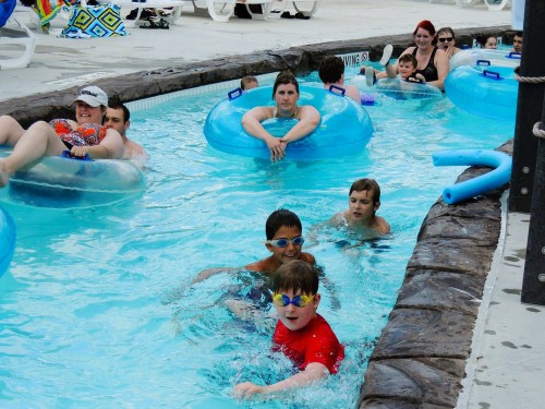 A group of children and adults relax in a lazy river pool, some on inner tubes, enjoying a sunny day.