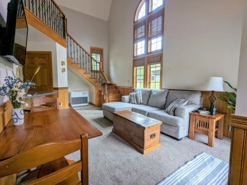 Cozy living room with a gray sofa, wooden furniture, and large windows, featuring a staircase in the background.