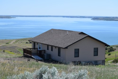 A house on a hillside overlooking a calm lake, surrounded by green grass and distant hills under a clear blue sky.