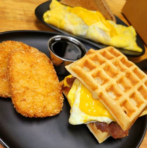 A plate with a waffle sandwich containing egg and cheese, alongside hash browns and a small cup of syrup.