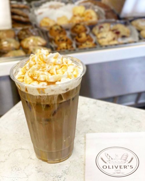 A frosty iced coffee topped with whipped cream and caramel, with baked goods in the background at Oliver's Café.