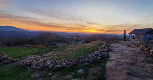 A person stands on a rocky path overlooking a sunset with rolling hills and a rustic building in the background.