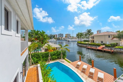 A sunny waterfront view featuring a pool, palm trees, and yachts along a canal, with blue skies and fluffy clouds.