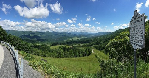 Panoramic view of green mountains and valleys under a blue sky with fluffy clouds, featuring a historical marker.