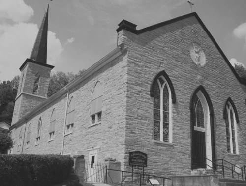 A historic stone church with a tall spire, arched windows, and a sign in front, set against a cloudy sky.