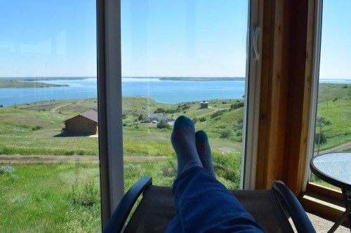 A person relaxes with feet up, overlooking a serene lake and green hills through a large window.