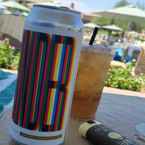 A colorful beer can next to a glass of iced tea and a sunscreen stick, set by a poolside with lounge chairs.