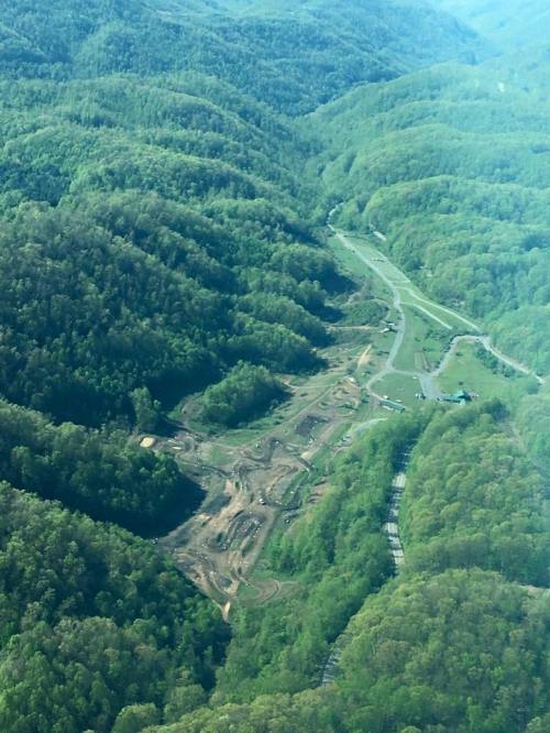 Aerial view of a green mountainous landscape with cleared land and construction areas visible among the trees.