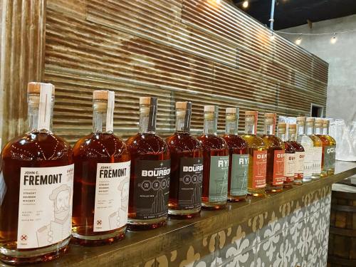 A row of various whiskey bottles on a wooden bar, featuring distinct labels and colors, set against a rustic backdrop.