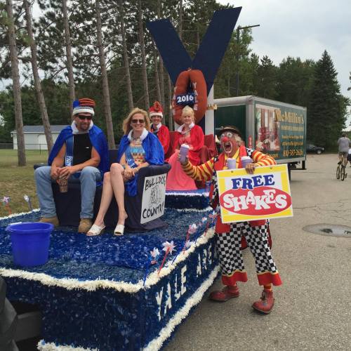 A festive parade float with people in costumes, including a clown offering free shakes, surrounded by trees.