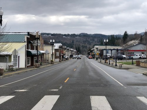 A quiet main street in a small town, lined with shops and trees, under a cloudy sky with mountains in the background.