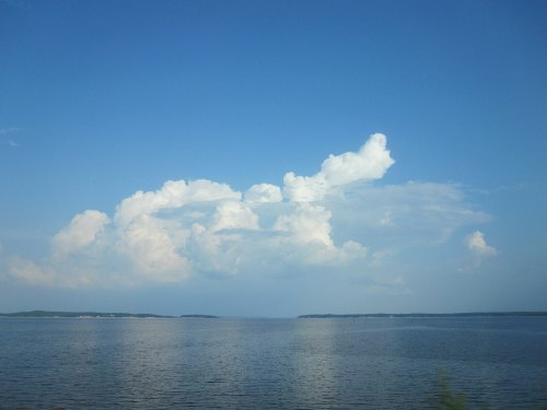 A serene lake scene with fluffy white clouds against a bright blue sky, reflecting on the calm water surface.
