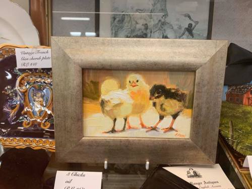 A framed painting of three chicks, two yellow and one black, displayed in an antique shop.