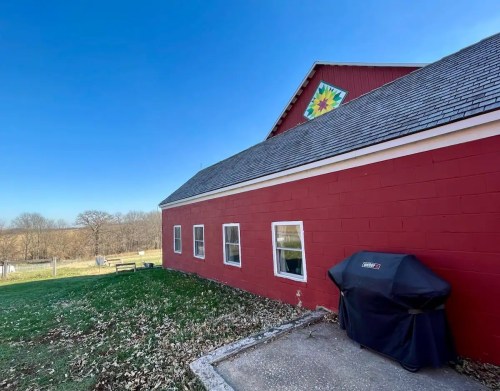 A red barn with a decorative quilt pattern on the roof, featuring a grill and a grassy area in the foreground.