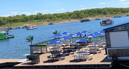 A lakeside dock with picnic tables and blue umbrellas, surrounded by water and green hills under a clear sky.
