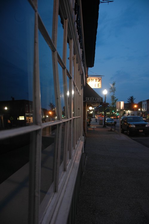 A quiet street at dusk, featuring storefronts and reflections in windows, with streetlights illuminating the scene.