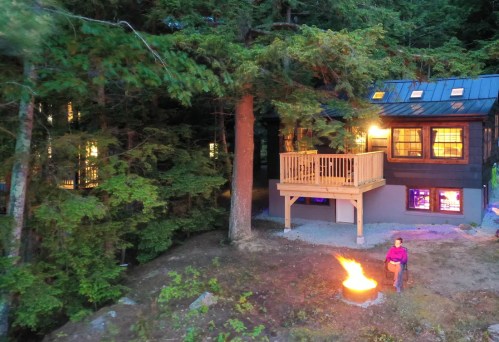 A cozy cabin in the woods with a fire pit, surrounded by tall trees and a person standing near the flames.