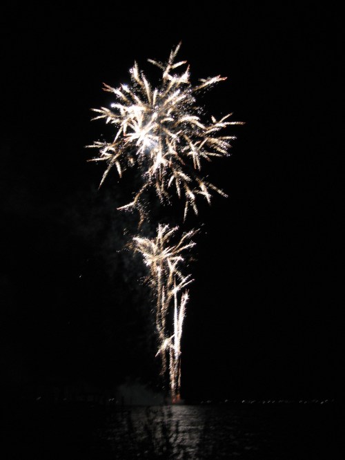 Colorful fireworks burst in the night sky over a dark body of water, creating a festive atmosphere.
