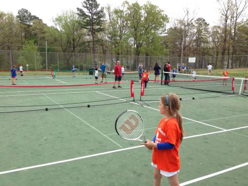 A young girl in an orange shirt holds a tennis racket, watching players on multiple courts in a park setting.