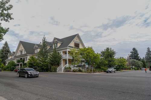 A large, multi-story house with a steep roof, surrounded by trees, at a street intersection on a cloudy day.