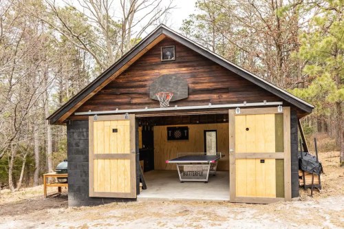 A rustic garage with wooden doors, a basketball hoop, and a ping pong table inside, surrounded by trees.
