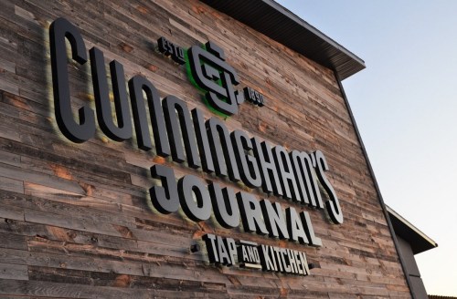 Sign for Cunningham's Journal Tap and Kitchen on a wooden wall, featuring a modern logo and established date.