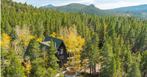 Aerial view of a cabin surrounded by lush green trees and mountains in the background, showcasing autumn colors.