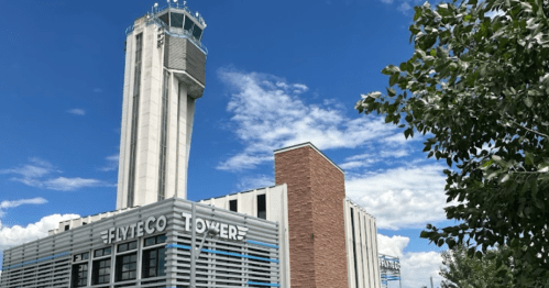 A tall tower with a glass observation deck, surrounded by a modern building and blue sky with clouds.