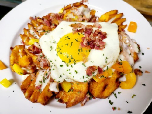A plate of waffle fries topped with a sunny-side-up egg, bacon bits, and diced peppers, garnished with herbs.