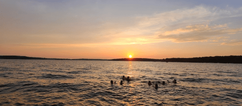 A serene sunset over a lake, with silhouettes of people swimming in the water.