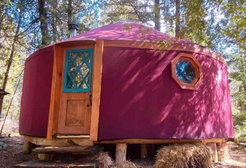 A colorful yurt with a wooden door and octagonal window, surrounded by trees and straw in a forest setting.