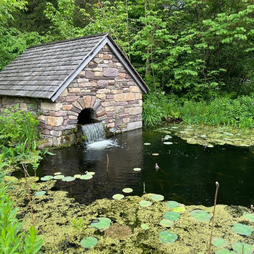 A stone building with a sloped roof beside a pond, featuring a small waterfall and surrounded by lush greenery and lily pads.