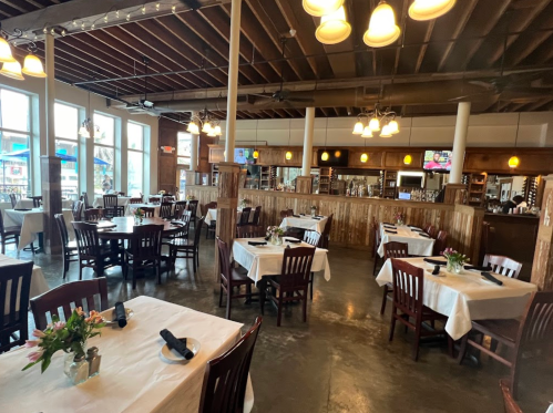 A spacious restaurant interior with wooden tables, chairs, and a warm ambiance, featuring flowers on each table.
