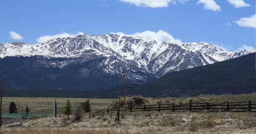 Snow-capped mountains under a blue sky with scattered clouds, framed by a grassy foreground and wooden fence.
