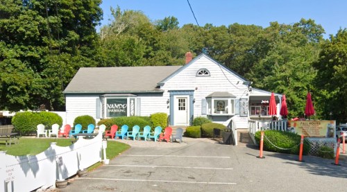 A quaint white building with a sign, surrounded by colorful chairs and trees, featuring outdoor seating and umbrellas.