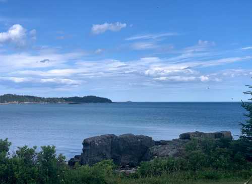 A serene coastal view featuring calm blue waters, distant islands, and a clear sky with scattered clouds.