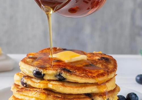 A stack of pancakes topped with butter and drizzled with syrup, with blueberries visible in the pancakes.