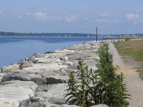 A rocky shoreline with a path beside calm water, under a clear blue sky with scattered clouds.