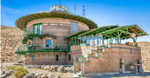 A unique round building with green accents, surrounded by rocky terrain and clear blue skies.