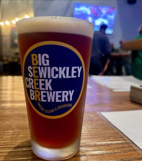 A pint glass of amber beer with a label for Big Sewickley Creek Brewery on a wooden table.