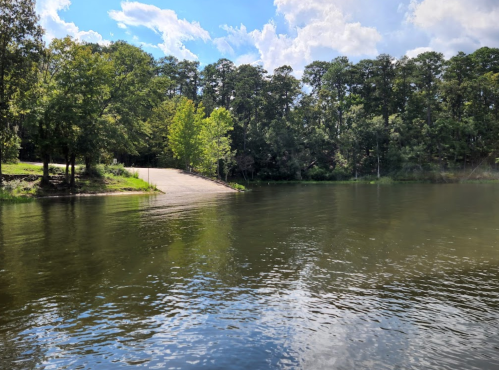 A serene lake scene with trees lining the shore and a small boat ramp leading to the water under a partly cloudy sky.