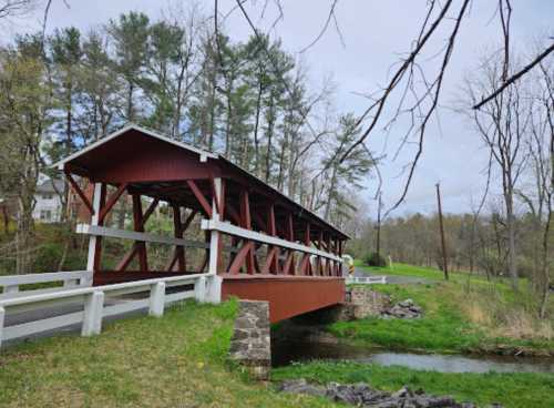 A red covered bridge spans a small stream, surrounded by trees and grassy areas on a cloudy day.